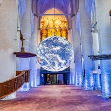 Foto des Innenraums der Hamburger Hauptkirche St. Katharinen. Zentral hängt die raumeinnehmende Installation "Gaia” - eine Erdkugel mit originalen NASA-Abbildungen und einem Durchmesser von sieben Metern. Die weißen Säulen werden blau angestrahlt.