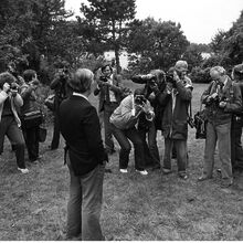 Schwarz-weiß Foto aus den 70er Jahren, auf dem Helmut Schmidt auf einer Wiese mit Blick auf einen See steht. Er ist von rund 12 Fotografen umringt. Das Foto ist hinter ihm aufgenommen worden.