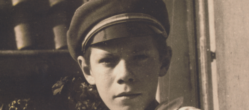 Portrait in sepia von Helmut Schmidt im Jugendalter. Er lehnt mit seinem rechten Ellenbogen an einem Blumenkasten. im Hintergrund ist ein Haus. Er trägt ein weißes Hemd und eine Art Kapitänsmütze. Sein Gesichtsausdruck ist neutral. Er schaut in die Kamera