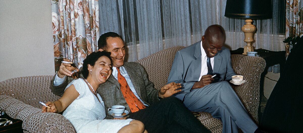 Foto, das wie ein privater Schnappschuss wirkt. Auf einem Sofa sitzen eine Frau und zwei Männer, die befreit lachen. Alle haben eine Zigarette in der Hand. Die Frau und ein Mann haben eine Kaffeetasse in der Hand, der andere Mann ein Schnapsglas.