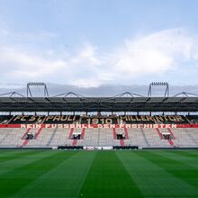 Foto einer überdachten Stadiontribüne. Davor ist ein akkurat gemähter Rasenfußballplatz. Auf der Tribüne steht: FC St. Pauli 1910 Millerntor. Kein Fußball den Faschisten.