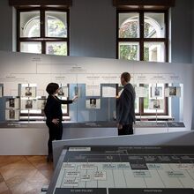Foto eines Ausstellungsbereichs. Eine Frau und ein Mann stehen vor einem breiten Exponat mit Stammbaumartigen Aufbau. An Stangen sind Tafeln mit je einem schwarz-weiß Portrait installiert, auf dessen Rückseite Text steht.