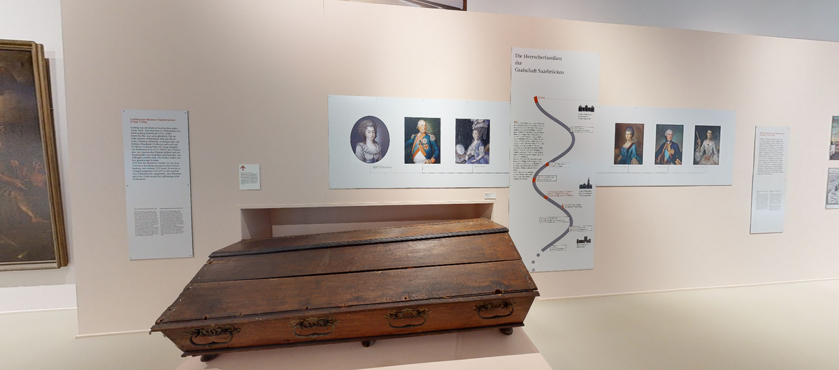 Foto eines Ausstellungsbereichs mit hellen Wänden und Boden. Auf einem flachen Podest steht ein älterer Sarg aus Holz. An der Wand dahinter hängen Informationstafeln mit Texten und älteren Portrait-Gemälden von Frauen und Männern.