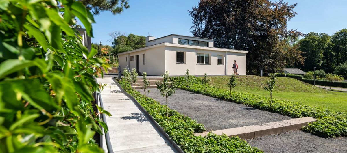 Foto des Haus am Horn. Zu dem weißen bungalowartigen Gebäude mit kleinen Aufbau führen ein Kiesweg, der von wenigen Treppen unterbrochen wird., sowie ein barrierefreier betonierter Weg. Drumherum sind grüne Hecken, Beete, Bäume und eine Rasenfläche.