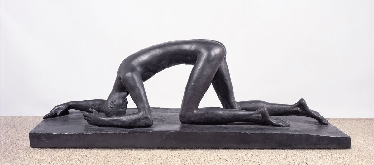Foto einer Bronze-Figur in einheitlichem Schwarz. Ein großer dünner nackter Mensch ist auf allen vieren auf einem niedrigen Sockel. Er schützt sich auf den Unterarmen und mit dem Kopf ab. Die Beine knien leicht versetzt.