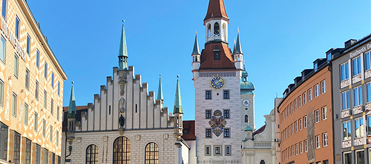 Foto des Marienplatzes in der Münchener Altstadt. Vor blauem Himmel ragen in der Mitte des Bildes ein heller Glockenturm mit einer großen und vier kleinen Turmspitzen sowie ein ebenfalls älteres weißes Gebäude mit vier Turmspitzen in den Himmel.