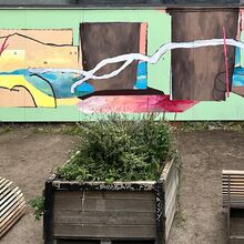Foto einer abstrakt bemalten Wand, die ca. 8 Meter lang und 2,5 Meter hoch ist. Davor stehen ein bepflanztes Hochbeet sowie rechts und links davon zwei Holzliegen. Der Boden besteht aus Sand und Erde.