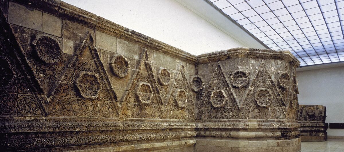 Hoher Raum mit originalgetreuer Mschatta-Fassade, die aus grauem Stein besteht, welcher mit hervorstehender Zickzack-Linie und steinernen Rosetten verziert ist.