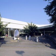 Foto des Eingangs vom Sprengel Museum Hannover. Langezogene flache Treppenstufen führen zu dem flachen Gebäude mit Glasfront und überdachtem Eingang. Eine abstrakte Skulptur steht auf den Stufen. Sieben Menschen halten sich auf den Stufen auf.