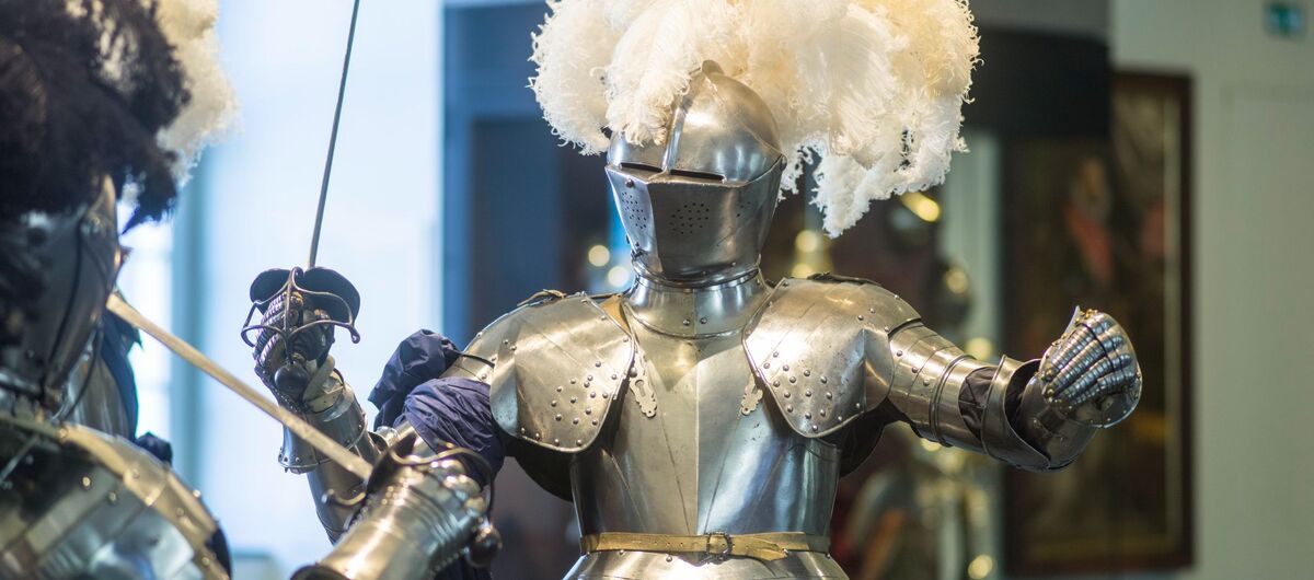 Foto von zwei miteinander kämpfenden Rittern in einem Ausstellungsraum. Die eine gut sichtbare Rüstung besteht zu den Metallelementen aus einem bestickten Samtstoff ab Hüfthöhe sowie ausladenden Federn am Helm.