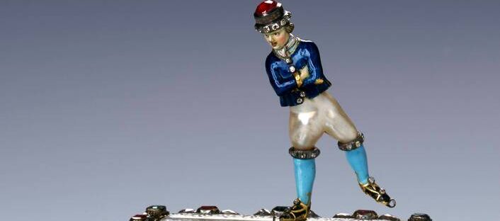 Foto einer Skulptur. Ein Schlittschuhläufer gleitet über die Eisfläche, die Arme vor der Brust verschränkt. Der kastenförmige, massive Sockel aus vergoldetem Silber ruht auf vier zierlichen Klauenfüßen. Die Seiten des Sockels zieren bunte Bilder.