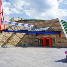 Außenansicht des Eingangsbereichs der Staatsgalerie Stuttgart. Das Sandsteingebäude wird durch moderne bunte Stahlelemente ergänzt. Die beiden zylinderförmigen Eingangselemente sind rot. Eine gebogene Fensterfront ist durch grüne Stahlträger unterbrochen.