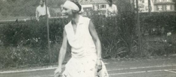 Schwarz-weiß Foto einer Frau in weißem Kleid und mit weißem Haarband mit Tennisschläger in ihrer rechten Hand. Sie steht im Ausfallschritt auf einem Tennisplatz und lächelt. Im Hintergrund stehen zwei Männer und schauen zu.