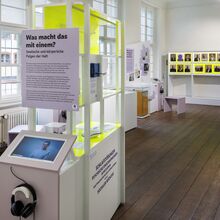 Foto eines Ausstellungsbereichs. Auf dunklem Holzboden stehen moderne Vermittlungsstationen in Form von Bildschirmen mit Kopfhörern, Vitrinen, Texttafeln sowie 19 kleine drehbare Tafeln mit Foto auf der Vorder- und Text auf der Rückseite.