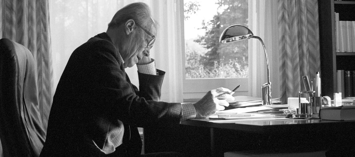 Schwarz-weiß Foto von Willy Brandt, der von der rechten Seite zu sehen an einem Schreibtisch sitzt. Er stützt seinen Kopf auf seinen linken auf den Schreibtisch aufgestützten Arm. Die rechte Hand liegt einen Stift haltend auf der Schreibtischplatte.