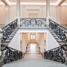 Ein feudales Treppenhaus mit Säulen und Marmor. Mittig geht eine Treppe hinunter, während links und rechts zwei Treppenaufgänge auf eine Empore führen. Das Geländer ist schwarz und sticht in dem hellen Ensemble, auch durch seine Verschnörkelungen hervor.