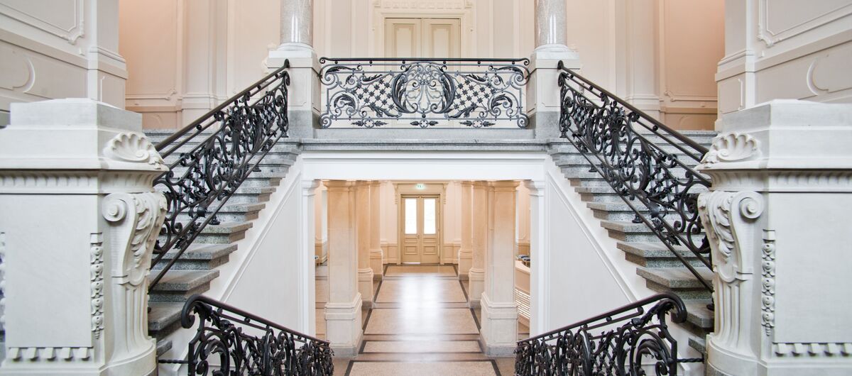 Ein feudales Treppenhaus mit Säulen und Marmor. Mittig geht eine Treppe hinunter, während links und rechts zwei Treppenaufgänge auf eine Empore führen. Das Geländer ist schwarz und sticht in dem hellen Ensemble, auch durch seine Verschnörkelungen hervor.