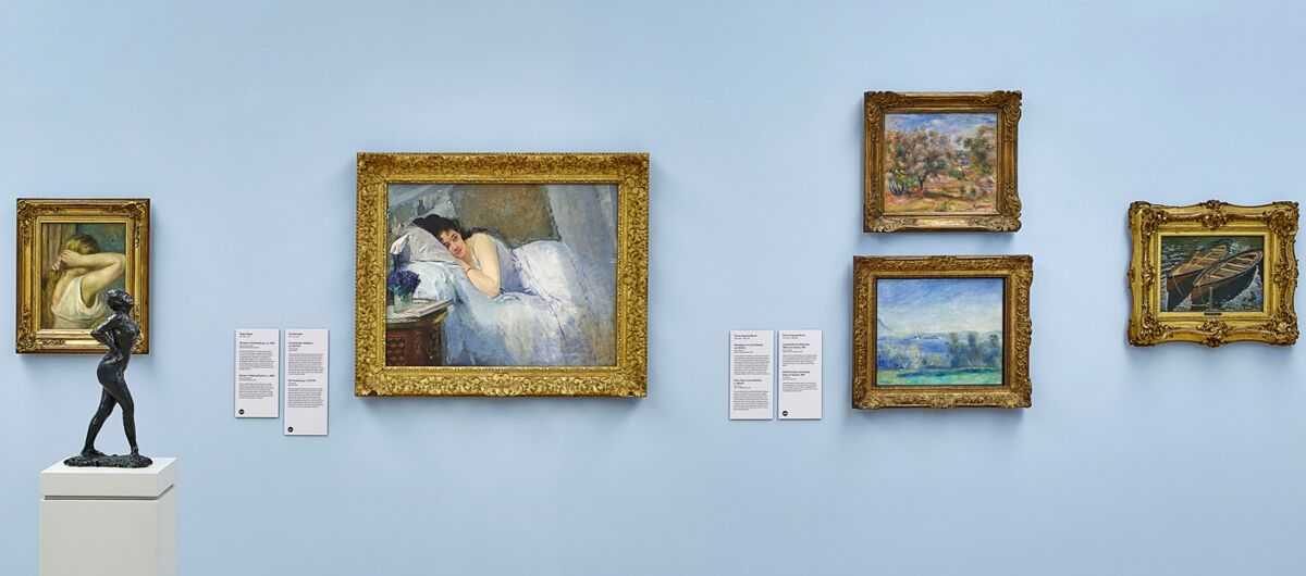 Hellblaue Wand eines Ausstellungsraumes, an der fünf unterschiedlich große Gemälde in prunkvollen Goldrahmen hängen.  Auf einem Sockel steht ein weiteres Kunstwerk: eine kleine Figur, die unbekleidet, schwarz gestaltet, ihre Hände ins Kreuz stemmt.