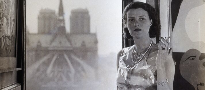 Schwarz-weiß Foto von Peggy Guggenheim. Sie steht im Kleid mit Perlenkette und Zigarette in ihrer linken gehobenen Hand vor einem Fenster in einem oberen Stockwerk. Ihre rchte Hand ruht auf einem Schrank vor dem Fenster.