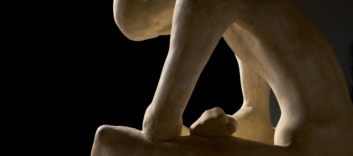 Foto einer hellen Skulptur vor schwarzem Hintergrund, die von rechts angestrahlt wird. Ein dünner nackter Mensch ohne Haare sitzt mit den Armen auf die Beine gestützt auf einem Stein. Man sieht ihn von seiner rechten Seite.