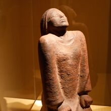 Foto einer Skulptur in einer Glasvitrine. Ein Mensch kniet in einem langen Gewand auf dem Boden. Die Hände liegen zu Fäusten geballt auf den Oberschenkeln. Die Augen sind geschlossen, der Kopf ist nach hinten gebeugt. Der Gesichtsausdruck ist neutral.