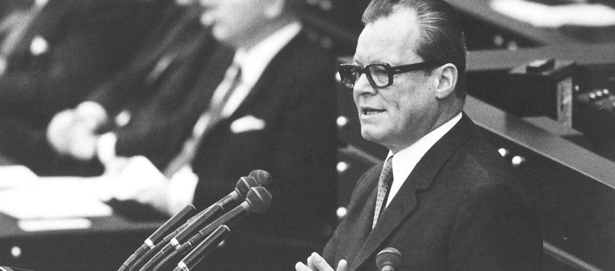 Schwarz-weiß Foto von Willy Brandt während einer Rede. Er steht an einem Pult mit sieben Mikrophonen, trägt einen schwarzen Anzug mit weißem Hemd und Krawatte sowie eine schwarze Brille mit dickem Rand. Seine Hände sind leicht gefaltet.