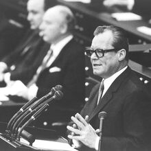 Schwarz-weiß Foto von Willy Brandt während einer Rede. Er steht an einem Pult mit sieben Mikrophonen, trägt einen schwarzen Anzug mit weißem Hemd und Krawatte sowie eine schwarze Brille mit dickem Rand. Seine Hände sind leicht gefaltet.