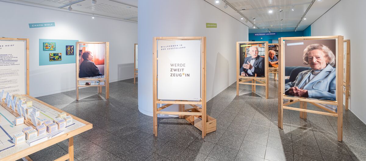 Foto eines Ausstellungsbereichs im Gustav-Lübcke Museum Hamm. Auf verschiedenen Holzaufstellern sind große Fotos von älteren Menschen zu sehen. Mittig steh ein Aufsteller mit den Worten "Werde Zweit Zeug*in".
