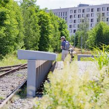 Foto mit Blick aus Kniehöhe. Links sind zwei Gleise zu sehen. Mittig eine lange Reihe von Informationstafeln aus Beton, die von einem Menschen mit Fahrrade betrachtet werden. Im Hintergrund sind grüne Bäume sowie eine Reihenhauszeile zu sehen.