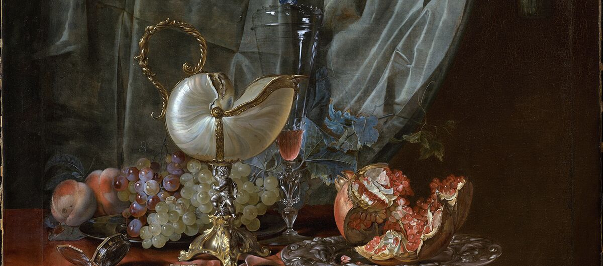 Stillleben mit Nautiluspokal, Pfirsichen, dunklen und grünen Trauben sowie einem auseinandergenommenem Granatapfel. auf einem Silbertablett. Das Stelleben steht auf einem roten Tuch. Dahinter hängt ein grauer Vorhang.