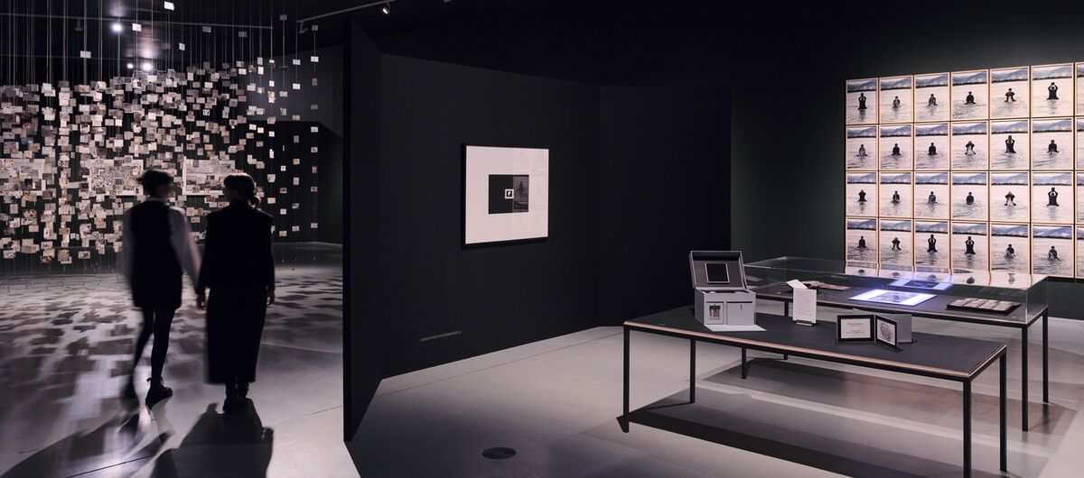 Schwarz-weiß Foto eines Ausstellungsbereichs. Es liegen Exponate auf zwei Tischen. An den beiden schwarzen Wänden drumherum sind ein einzelnes sowie eine Fotoreihe mit über 24 Fotos zu sehen. Viele kleine Zettel hängen links von der Decke.