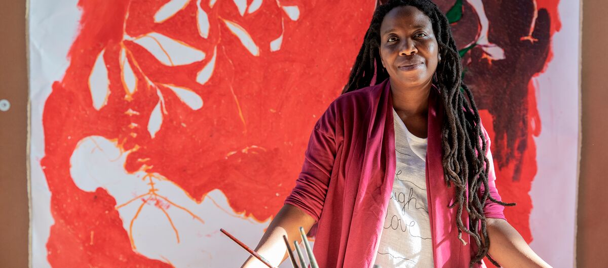 Das Foto zeigt die Künstlerin Portia Zvavahera in ihrem Studio. Sie ist eine schwarze Frau mit Dreadlocks bis zum Bauch. Ihr Blick ist freundlich. Sie stützt sich an ihrem Tisch ab, auf denen Pinsel und Farbdosen stehen. Im Hintergrund ein Gemälde.