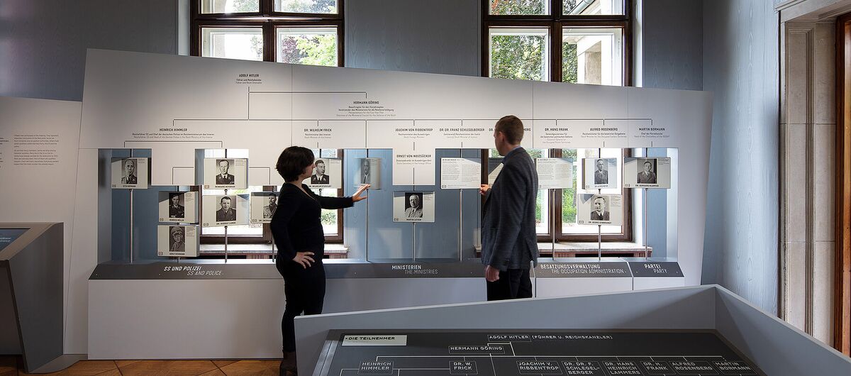 Foto eines Ausstellungsbereichs. Eine Frau und ein Mann stehen vor einem breiten Exponat mit Stammbaumartigen Aufbau. An Stangen sind Tafeln mit je einem schwarz-weiß Portrait installiert, auf dessen Rückseite Text steht.