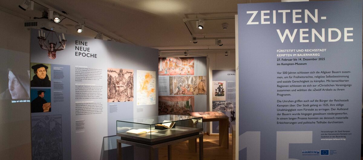 Foto eines Teils der Ausstellung Zeitenwende. In einem dunklen Raum stehen in blau gehaltene Informationswände sowie Vitrinen, die durch Lichtspotts angestrahlt werden. Auf eine Wand wird etwas projiziert.