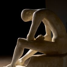 Foto einer hellen Skulptur vor schwarzem Hintergrund, die von rechts angestrahlt wird. Ein dünner nackter Mensch ohne Haare sitzt mit den Armen auf die Beine gestützt auf einem Stein. Man sieht ihn von seiner rechten Seite.