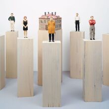 Foto von sieben Figuren und einem Haus aus bemaltem Holz, die jeweils auf einem Holzsockel stehen. Die Figuren stehen, haben aber unterschiedliche Körperhaltungen. Das dreigeschossige Haus ist würfelartig mit vier Schornsteinen.