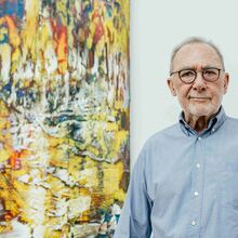 Gerhard Richter mit grauem Haar, Brille und hellblauem Hemd steht vor einem großformatigen, farbenfrohen abstrakten Gemälde. Das Bild im Hintergrund zeigt eine lebendige Mischung aus Gelb-, Rot- und Blautönen.