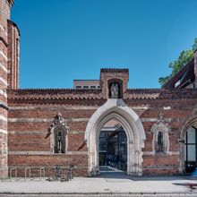 Foto von St. Annen bei Sonnenschein. Der Backsteinbau ist durch waagerechte Sandsteinlinien durchzogen. Die Mauer ist durch einen Durchgang unterbrochen. Rechts und links daneben ist je eine Einbuchtung mit einer Figur zu sehen.