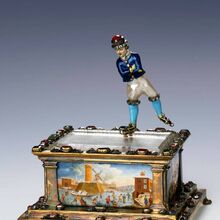 Foto einer Skulptur. Ein Schlittschuhläufer gleitet über die Eisfläche, die Arme vor der Brust verschränkt. Der kastenförmige, massive Sockel aus vergoldetem Silber ruht auf vier zierlichen Klauenfüßen. Die Seiten des Sockels zieren bunte Bilder.