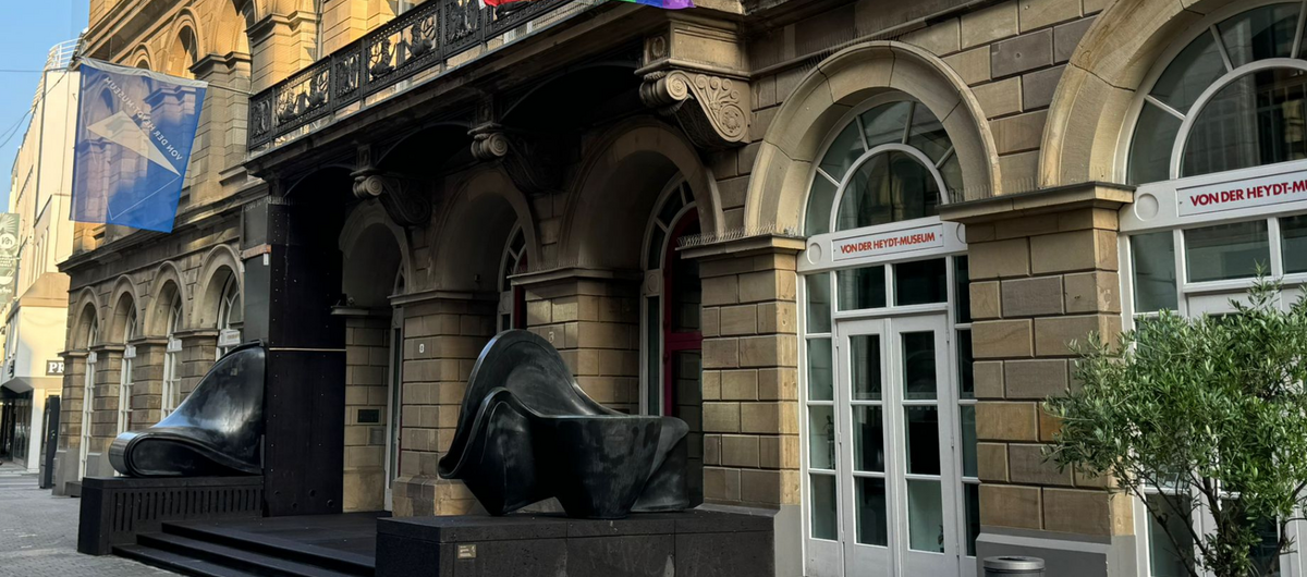 Foto der Außenansicht des Von der Heydt-Museums. Zu sehen ist der Eingangsbereich, der durch zwei abstrakte schwarze Skulpturen gesäumt wird. Auf höhe der ersten Etage hängt eine Fahne des Museums sowie eine Regenbogenfahne.
