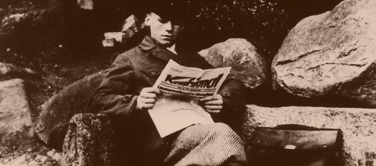 Schwarz-weiß Foto eines jungen gut gekleideten Mannes, der mit überschlagenen Beinen auf einer Bank aus großen Steinen sitzt und Zeitung liest. Neben ihm auf der Bank liegt eine Aktentasche.