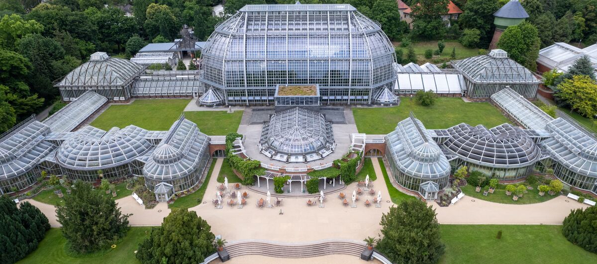 Luftaufnahme der Gebäude des Botanischen Gartens Berlin. Zentral steht ein kuppelartiges großes Glashaus. Davor rechteckig angeordnet stehen neun kleinere Glashäuser in unterschiedlichen Formen, die durch kurze Gänge miteinander verbunden sind.