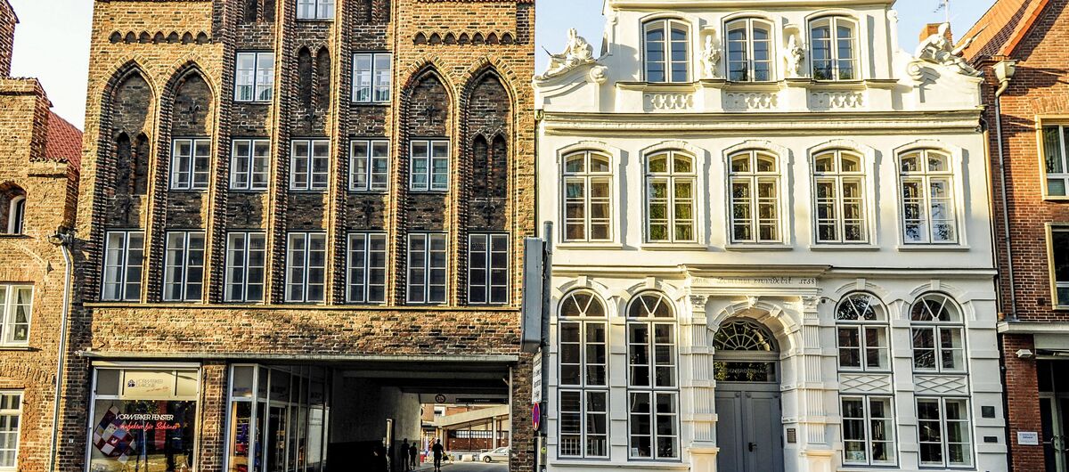 Foto einer historischen Häuserreihe. Auf der rechten Seite des Fotos ist mit weißer Fassade das dreistöckige Buddenbrookhaus zu sehen. Die linke Seite wird durch einen ähnlich hohen Klinkerbau mit großem Durchgang zum Hinterhof dominiert.