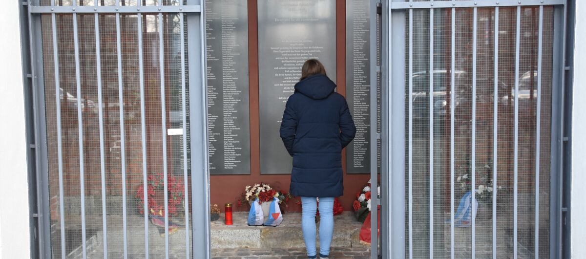 Eine Frau vor Gedenktafeln, auf denen unzählige Namen stehen. Die Tafeln sind in einem Raum, der hinter Gittern liegt und über eine geöffnete Tür betretbar ist. Vor den Gedenktafeln stehen auf dem Boden Grabkerzen und Kränze liegen nieder.