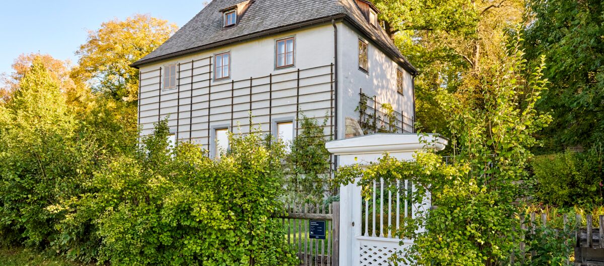 Weißes Haus in grünem Garten, zweigeschossig mit Schiefergedecktem Spitzdach. An den Wänden leere Rankhilfen, die Sprossenfenster sind mit Gardinen verhangen. Ein Zaun mit weißem Gartentor begrenzt das Grundstück, welches in warmen Sonnenlicht liegt.