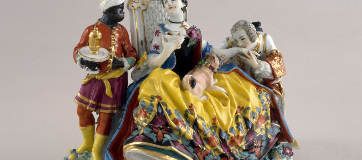 Aus Meissner Porzelan gefertigste Szenerie: Eine Dame sitzt auf einem Stuhl und hält eine Tasse in ihrer rechten. Auf ihrem Schoss ein Hündchen. Den linken Arm streckt sie einer Figur zur Kusshand hin. Rechts ein Bediensteter mit dunkler Hautfarbe