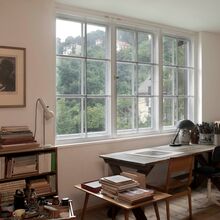 Foto eines Arbeitszimmers mit Schreibtisch und zwei Stühlen vor einem großen Sprossenfenster mit Blick auf einen bewaldeten Berg. Im Raum stehen weitere Schränke und Tische aus Holz mit Büchern und Zeichen- bzw. Schreibutensilien.