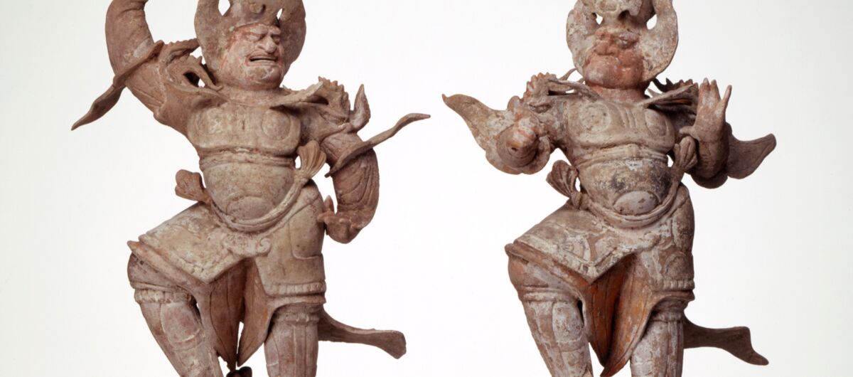 Abbildung zweier Holzfiguren aus China. Sie sind kunstvoll geschnitzt, unbemalt. Beide tragen eine Art Rüstung und Kopfschmuck, sie befinden sich in Bewegung, mit teils erhobener Faust und wehenden Capes und Bändern. Ihr rechter Fuß ruht auf einem Tier.