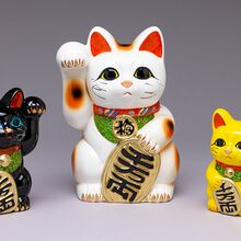 Auf dem Bild sind drei Maneki-Neko, die japanischen Glückskatzen, zu sehen. Sie unterscheiden sich in Größe und Farbe. Jede Katze hat ein freundliches Gesicht und hält eine Pfote erhoben, wie es typisch für Maneki-Neko ist.
