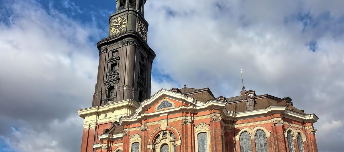 Seitlicher Blick auf die Michaeliskirche (Michel) mit Kirchenschiff in rotem Stein, geschwungenen und hell eingefassten Fenstereinfassungen sowie Dachsimsen. Links der hohe Turm, der aus dunklem Material besteht und in den blauen Himmel ragt.